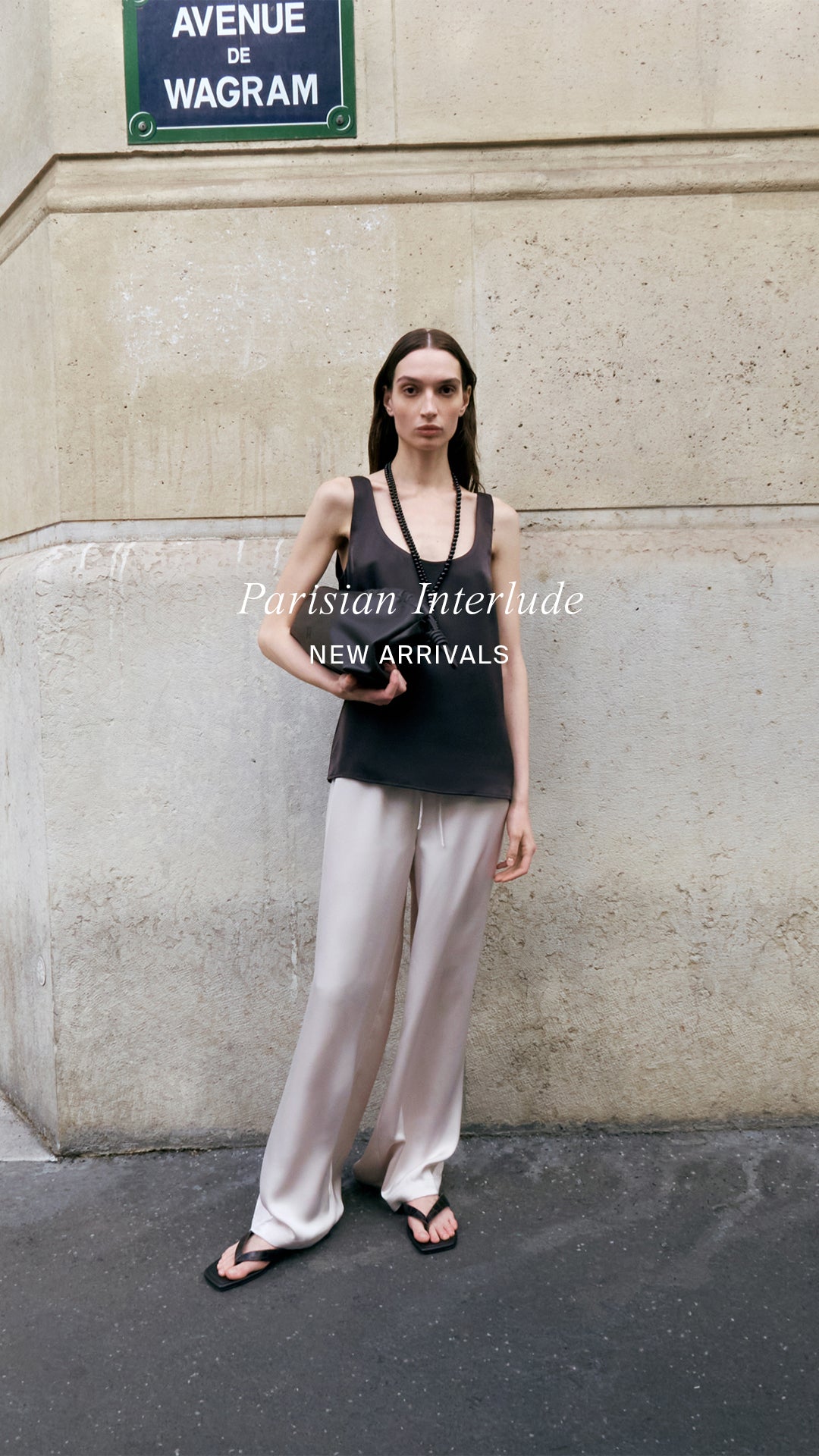 Woman standing against a wall with a street sign in the background, wearing a brown tank top and light-colored pants.