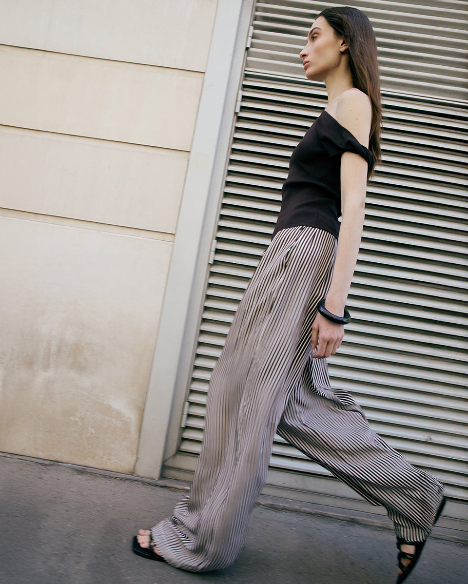 Woman wearing a brown top and striped pants walking past a building with horizontal slats.