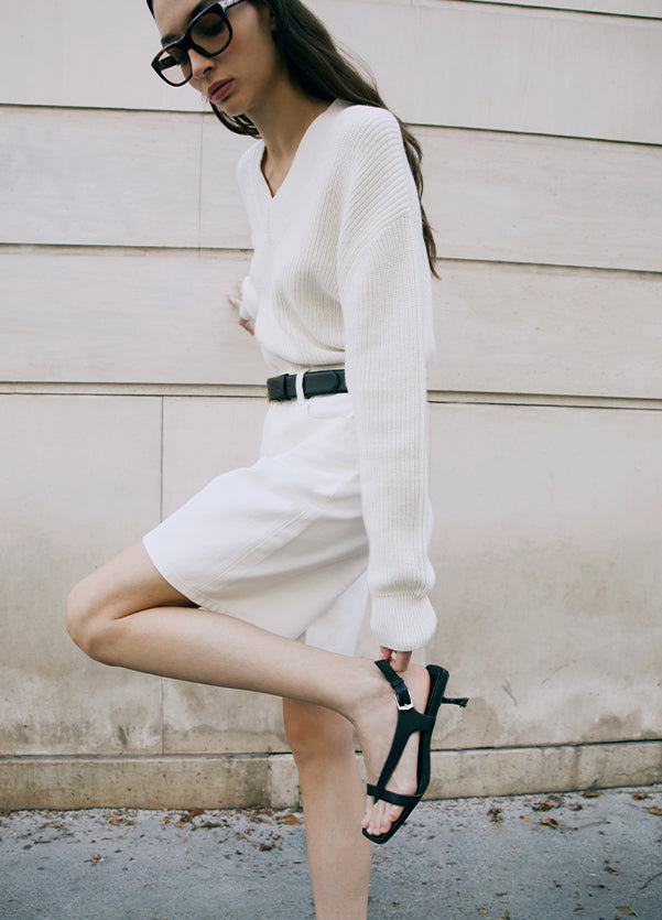 Woman in a white outfit with black high-heeled shoes against a beige wall.
