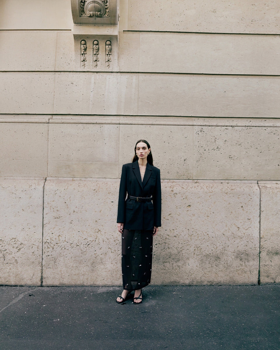 Woman in a black suit standing against a textured wall.